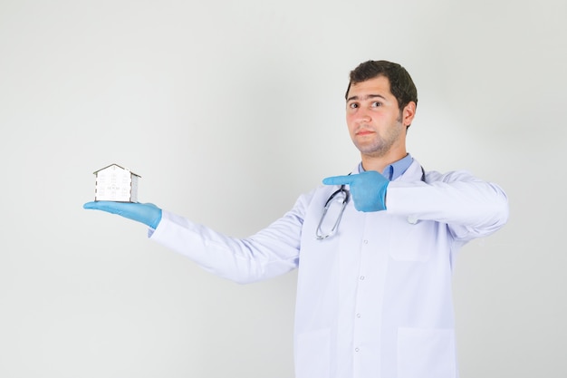 Doctor de sexo masculino que señala el dedo en el modelo de la casa en bata blanca, vista frontal de los guantes.