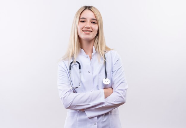 Doctor sonriente joven rubia con estetoscopio y bata médica en corsé dental cruzando las manos sobre fondo blanco aislado