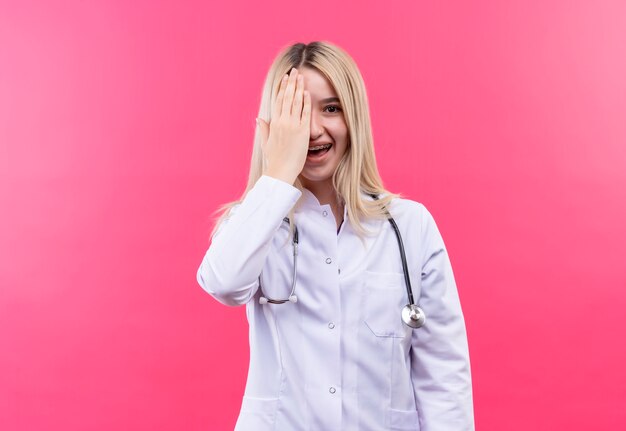 Doctor sonriente joven rubia con estetoscopio en bata médica y corsé dental cubierto ojo sobre fondo rosa aislado