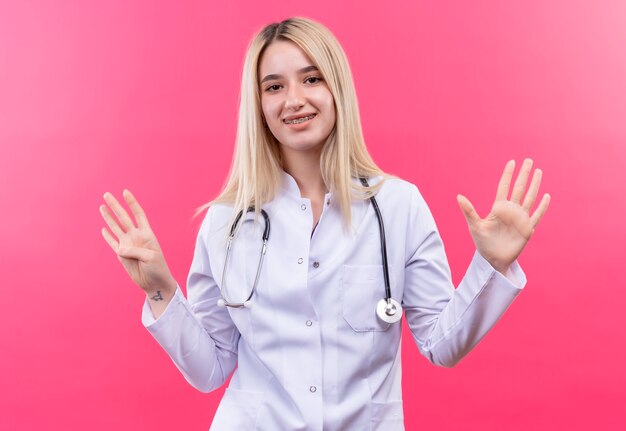 Doctor sonriente joven rubia con estetoscopio en bata médica y corsé dental mostrando diferentes números con las manos sobre fondo rosa aislado