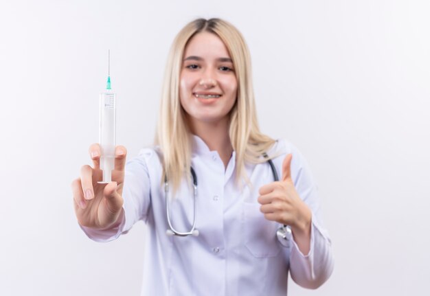 Doctor sonriente joven rubia con estetoscopio y bata médica en corsé dental sosteniendo la jeringa con el pulgar hacia arriba sobre fondo blanco aislado