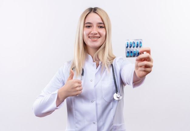 Doctor sonriente joven rubia con estetoscopio y bata médica en corsé dental sosteniendo pastillas con el pulgar hacia arriba sobre fondo blanco aislado