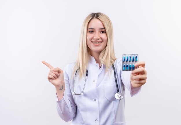 Doctor sonriente joven rubia con estetoscopio y bata médica en corsé dental sosteniendo píldoras apunta a un lado sobre fondo blanco aislado