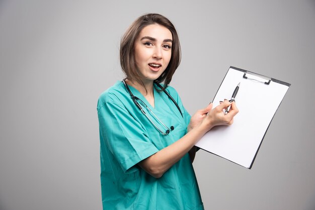 Doctor sorprendido apuntando al portapapeles en la pared gris.