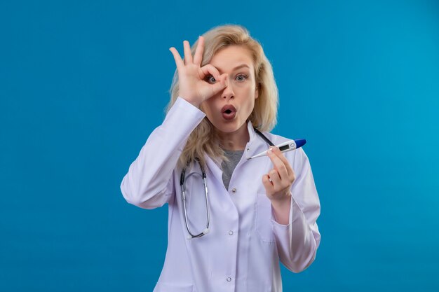 Doctor sorprendido joven con estetoscopio en bata médica sosteniendo un termómetro mostrando gesto de mirada sobre fondo azul.