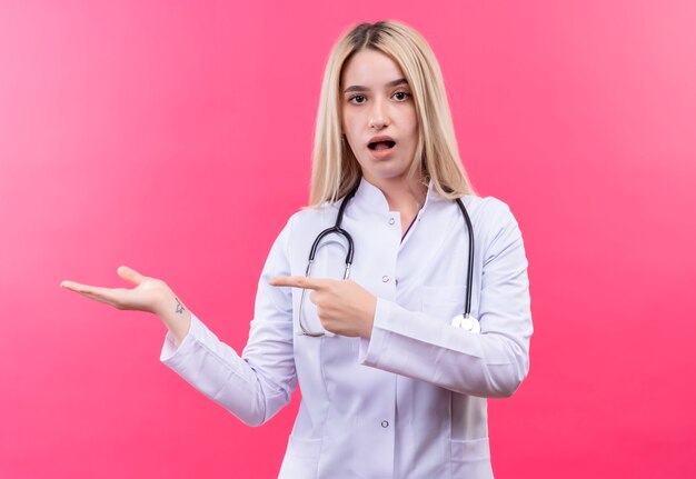 Doctor sorprendido joven rubia con estetoscopio en bata médica apunta al lado sobre fondo rosa aislado