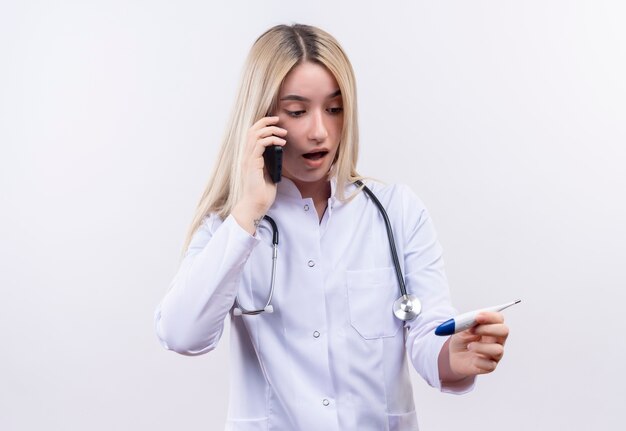 Doctor sorprendido joven rubia con estetoscopio en bata médica habla por teléfono mirando el termómetro en su mano sobre fondo blanco aislado