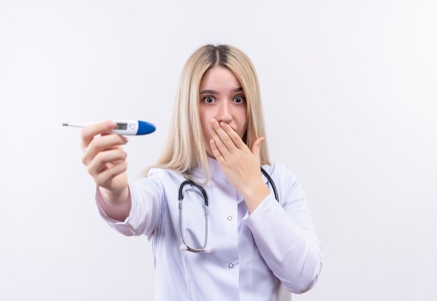 Doctor sorprendido joven rubia con estetoscopio y bata médica sosteniendo el termómetro a la cámara con la boca cubierta sobre fondo blanco aislado