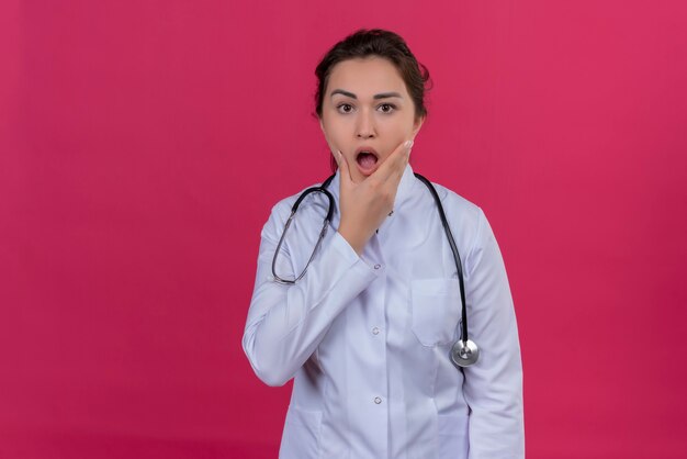 Doctor sorprendido joven vistiendo bata médica y estetoscopio agarró la mandíbula sobre fondo rojo isoleted