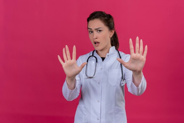 Doctor sorprendido joven vistiendo bata médica y estetoscopio mostrando gesto de parada sobre fondo rojo isoleted