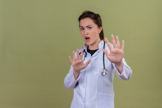 Doctor sorprendido joven vistiendo bata médica con estetoscopio muestra gesto de parada sobre fondo verde