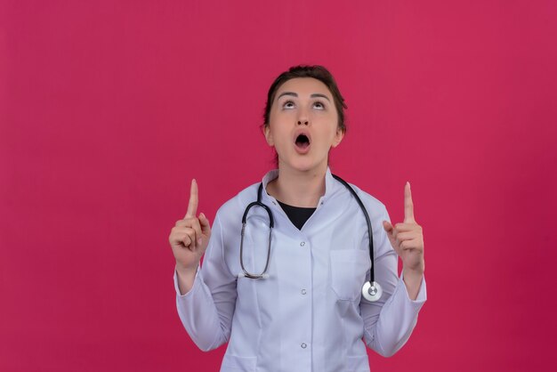 Doctor sorprendido joven vistiendo bata médica y puntos de estetoscopio hacia arriba sobre fondo rojo isoleted