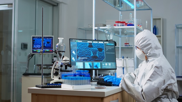 Doctor en traje de ppe mirando muestra de sangre en tubo de ensayo sentado en laboratorio equipado. Científico que trabaja con diversas bacterias y tejidos, concepto de investigación farmacéutica de antibióticos contra covid19