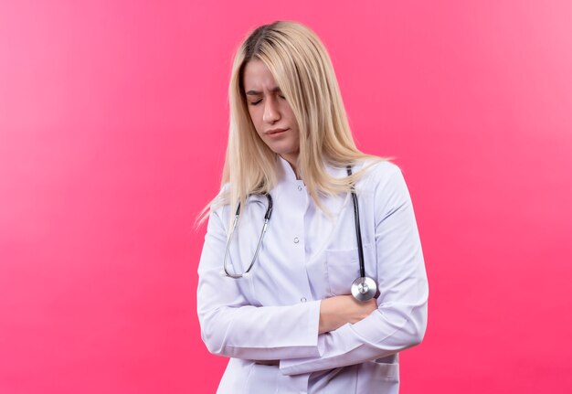 Doctor triste joven rubia con estetoscopio en bata médica cruzando las manos sobre fondo rosa aislado