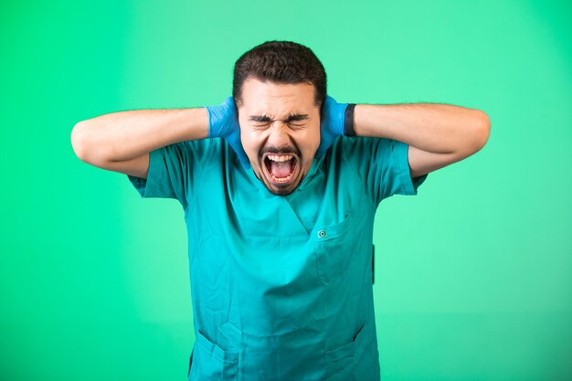 Doctor en uniforme y máscara de mano cubriendo sus oídos y con shock emocional, sobre fondo verde.