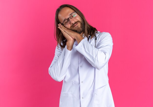 Doctor varón adulto complacido vistiendo bata médica y estetoscopio con gafas haciendo gesto de sueño con los ojos cerrados