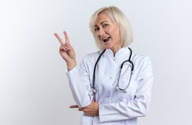 Doctora adulta alegre en bata médica con estetoscopio gesticulando el signo de la victoria aislado en la pared blanca con espacio de copia