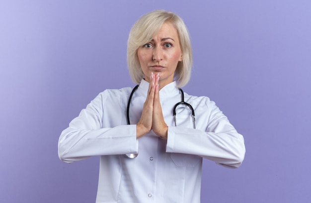 Doctora adulta ansiosa en bata médica con estetoscopio tomados de la mano juntos aislado en la pared púrpura con espacio de copia