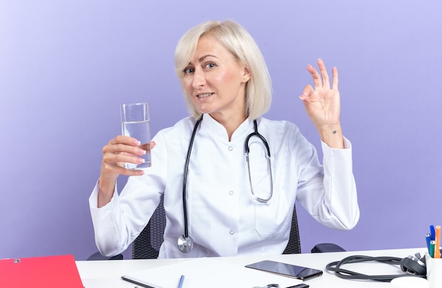 Doctora adulta complacida en bata médica con estetoscopio sentado en el escritorio con herramientas de oficina sosteniendo un vaso de agua y gesticulando el signo de ok aislado en la pared púrpura con espacio de copia