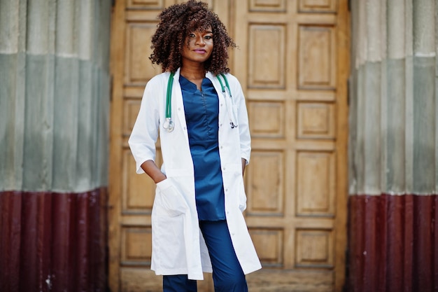 Doctora afroamericana en bata de laboratorio con estetoscopio al aire libre contra la puerta de la clínica