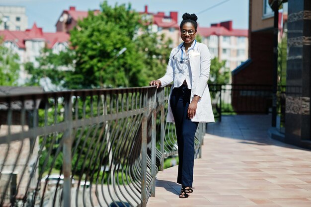 Doctora afroamericana en bata de laboratorio con estetoscopio posada al aire libre contra la clínica