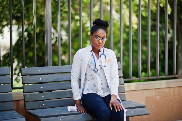 Doctora afroamericana en bata de laboratorio con estetoscopio posada al aire libre sentada en un banco