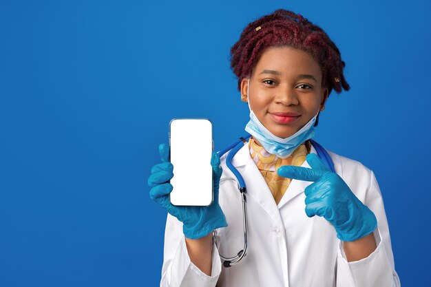 Doctora afroamericana mostrando la pantalla del teléfono inteligente en blanco sobre fondo azul.