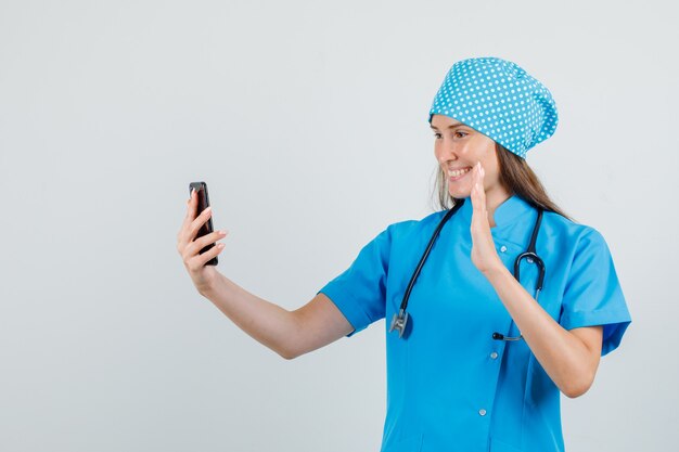 Doctora agitando la mano en la videollamada en uniforme azul y mirando alegre