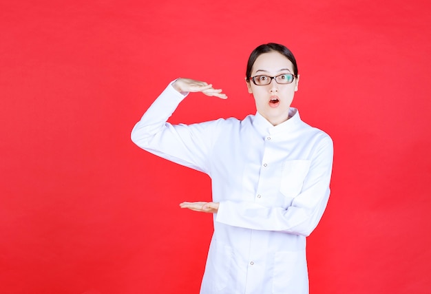 Doctora en anteojos de pie sobre fondo rojo y mostrando el tamaño de un objeto.