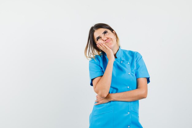 Doctora apoyando la mejilla en la palma levantada en uniforme azul y mirando vacilante