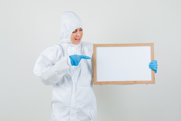 Doctora apuntando al marco en blanco con traje protector, guantes y mirando feliz.