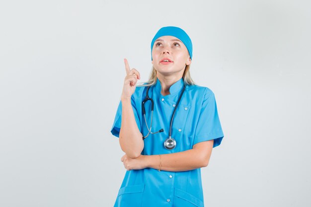 Doctora apuntando hacia arriba con el dedo en uniforme azul y mirando esperanzado