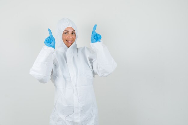 Doctora apuntando hacia arriba en traje de protección, guantes y mirando feliz