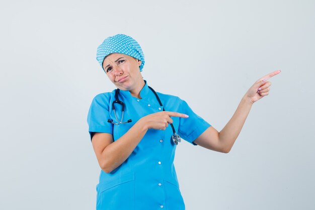 Doctora apuntando a la esquina superior derecha en uniforme azul y mirando decepcionada. vista frontal.