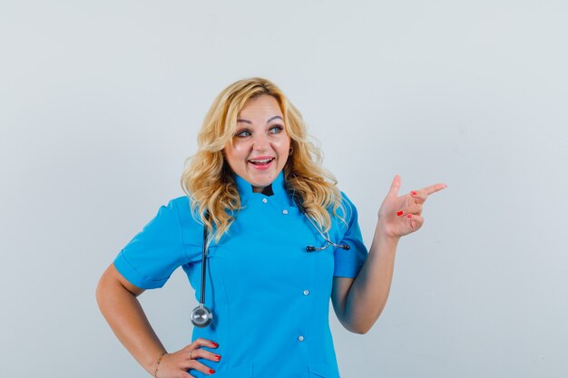 Doctora apuntando a un lado en uniforme azul y mirando contenta.