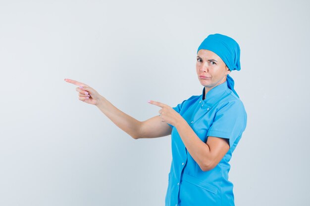 Doctora apuntando hacia el lado en uniforme azul y mirando dudoso, vista frontal.