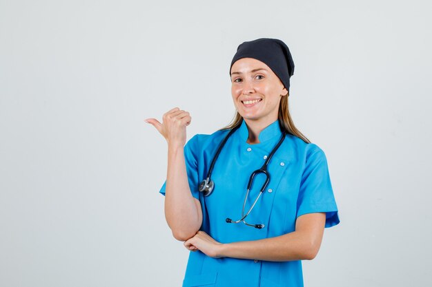 Doctora apuntando con el pulgar al lado en uniforme y mirando alegre