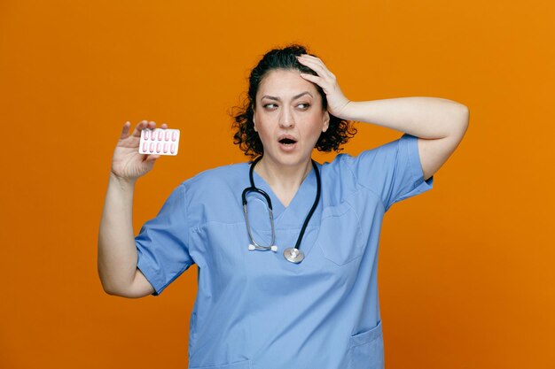 Una doctora arrepentida de mediana edad que usa uniforme y estetoscopio alrededor de su cuello muestra un paquete de cápsulas mirando a un lado mientras mantiene la mano en la cabeza aislada en un fondo naranja