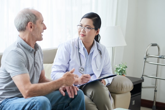 Doctora asiática hablando con paciente caucásico senior durante visita a domicilio