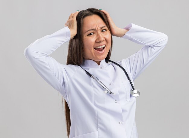 Doctora asiática joven estresada vistiendo bata médica y estetoscopio mirando al frente manteniendo las manos en la cabeza gritando aislado en la pared blanca