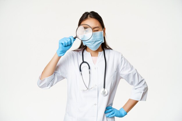 Doctora asiática mirando a través de una lupa, examinar al paciente, buscar enfermedades, usar mascarilla médica y guantes de goma, fondo blanco.