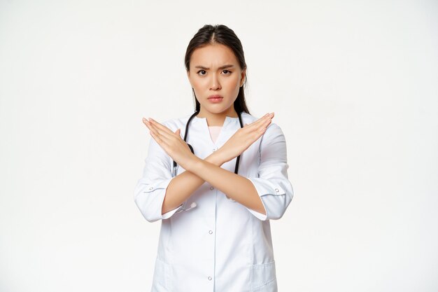 La doctora asiática seria prohíbe el comportamiento del paciente, muestra parada, gesto de brazos cruzados, frunce el ceño disgustado, desaprueba algo malo, de pie sobre fondo blanco