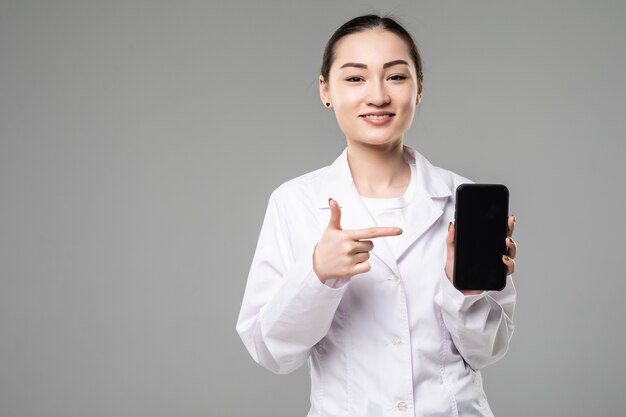 Doctora asiática sonriendo y mostrando una pantalla de teléfono inteligente en blanco aislada en una pared blanca