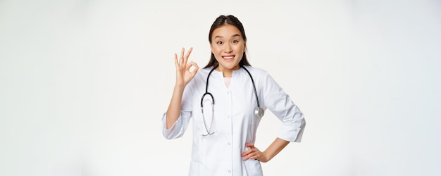 Una doctora asiática sonriente muestra un signo de estar bien y usa una bata médica una trabajadora del hospital en uniforme recomienda algo de pie sobre un fondo blanco
