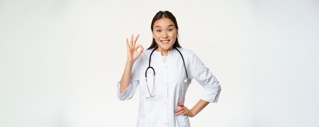 Una doctora asiática sonriente muestra un signo de estar bien y usa una bata médica Una trabajadora del hospital en uniforme recomienda algo de pie sobre un fondo blanco