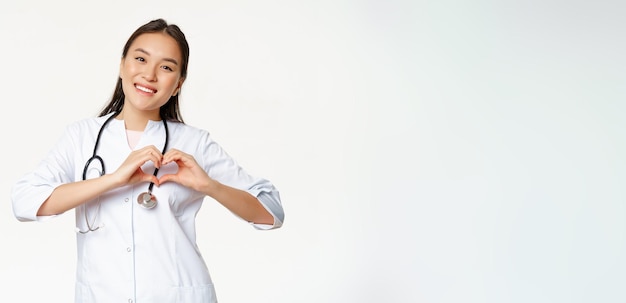 Foto gratuita una doctora asiática sonriente que atiende a sus pacientes muestra un gesto cardíaco y se ve feliz de pie