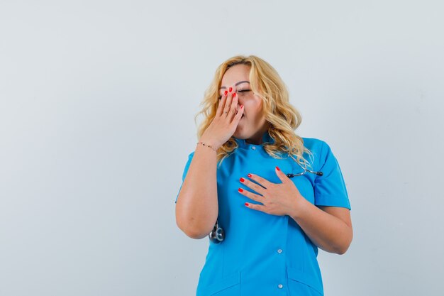 Doctora bostezando con la mano en la boca en uniforme azul y mirando espacio soñoliento para texto
