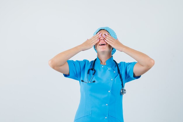 Doctora cogidos de la mano en los ojos en uniforme azul y mirando emocionado, vista frontal.