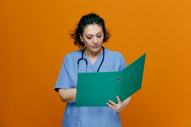 Doctora concentrada de mediana edad con uniforme y estetoscopio alrededor de su cuello sosteniendo una carpeta apuntando con la mano leyendo de ella aislada en un fondo naranja