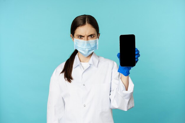 Doctora enojada y confundida con guantes de máscara facial que muestra la pantalla móvil de la aplicación del teléfono inteligente frunciendo el ceño...